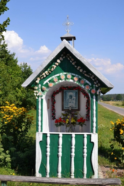 Mała kapliczka polowa w zielono-białych kolorach z dachem, ozdobiona kwiatami, z obrazem wewnątrz, ustawiona przy drodze na tle zieleni i błękitnego nieba.