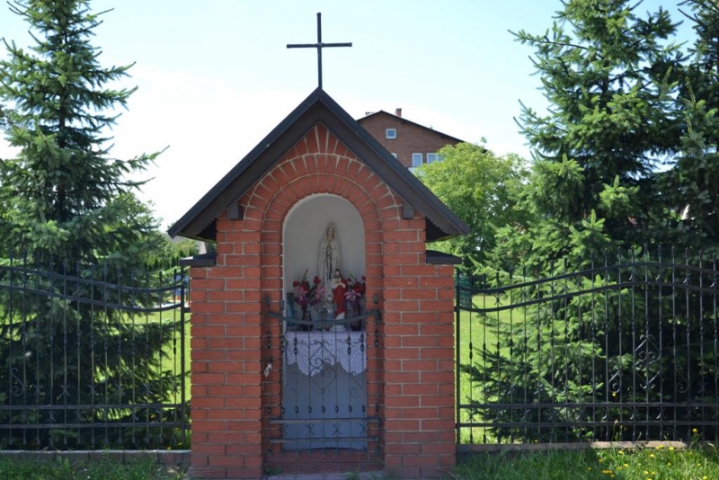 Kapliczka z czerwonej cegły z figurą Matki Boskiej, ozdobiona kwiatami, otoczona czarnym metalowym ogrodzeniem, na tle zielonych drzew i budynku.