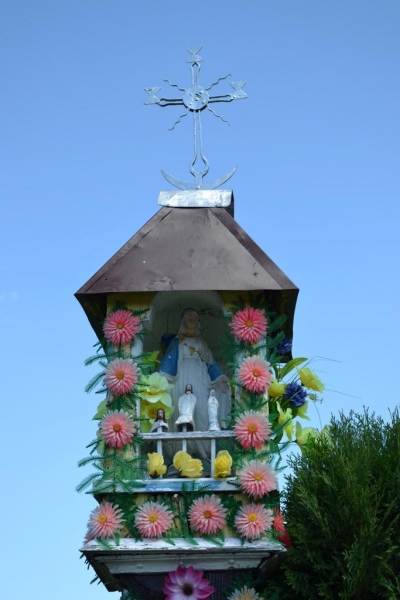 Kapliczka z brązowym dachem, ozdobiona różowymi i żółtymi sztucznymi kwiatami, z figurkami religijnymi w środku, na tle jasnoniebieskiego nieba.