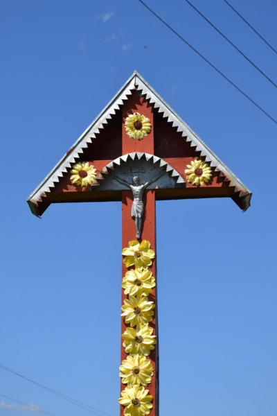 Czerwony drewniany krzyż z metalową figurą Jezusa, ozdobiony dużymi żółtymi kwiatami, na tle błękitnego nieba z kilkoma cienkimi chmurami i przewodami elektrycznymi.