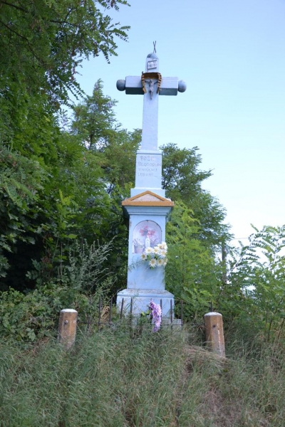 Kamienny biały krzyż z ozdobami i bukietami kwiatów na tle zielonych drzew i niebieskiego nieba.
