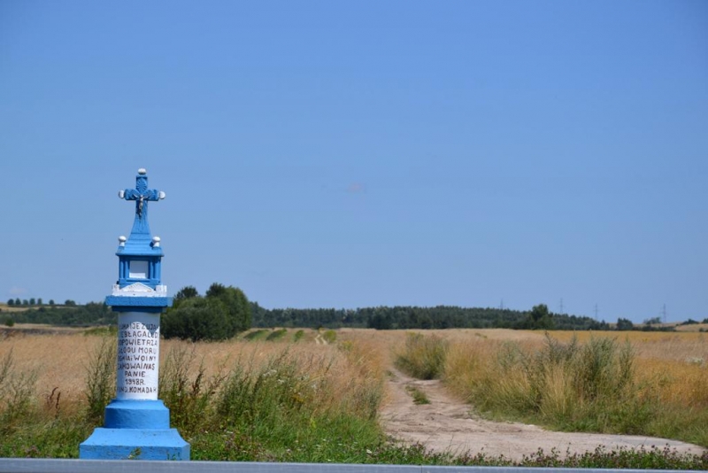 Niebieski drewniany krzyż z napisem na białej podstawie, sucha trawa, polna droga, zielone krzewy, błękitne niebo.
