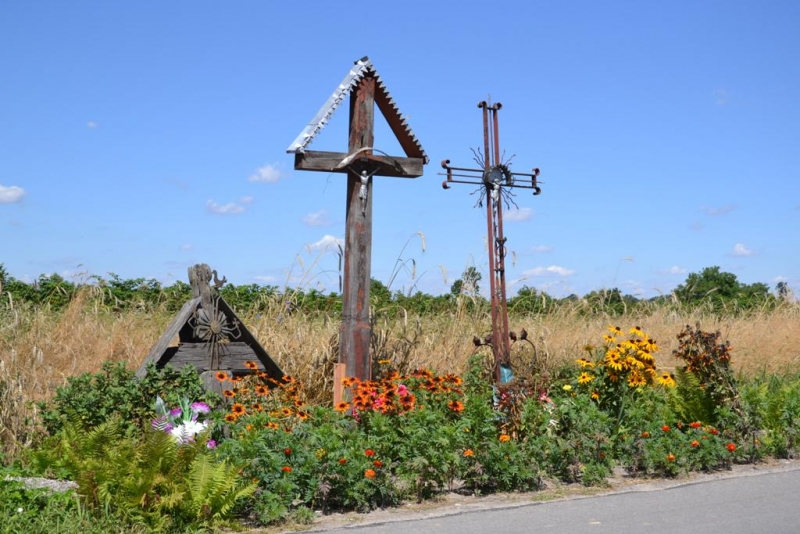Trzy drewniane i metalowe krzyże wśród zieleni i kolorowych kwiatów na tle suchej trawy i niebieskiego nieba.