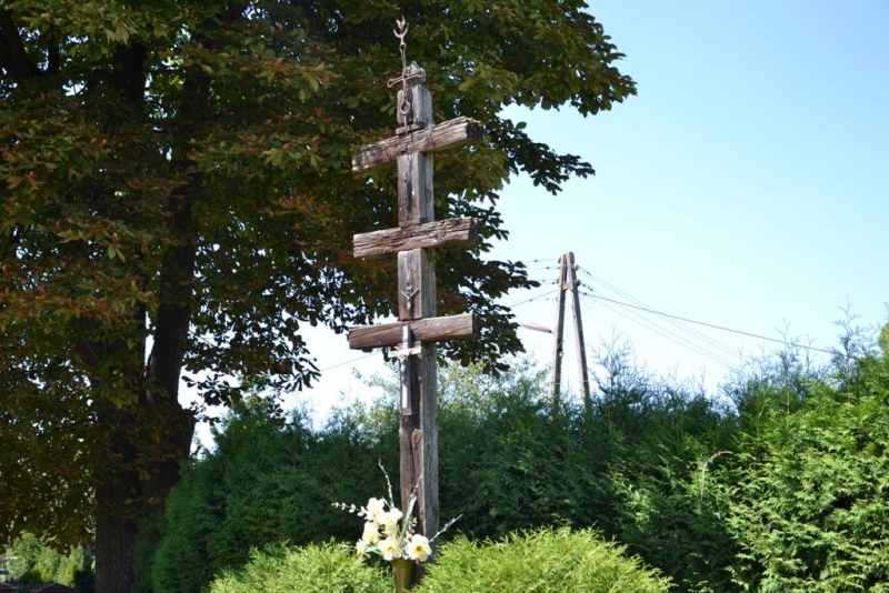 Stary drewniany krzyż z trzema poprzeczkami na tle zielonych drzew i krzewów, u dołu kwiaty, jasne niebo.