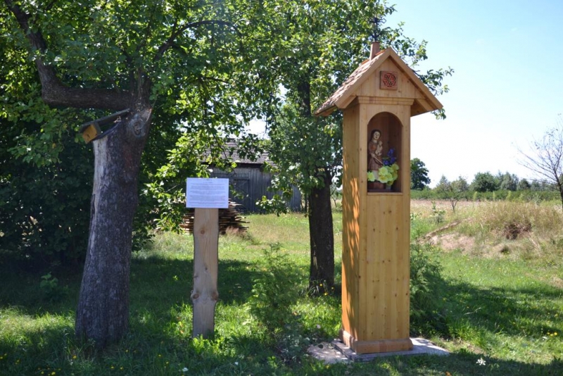 Drewniana kapliczka z figurką i kwiatami na zielonej polanie, obok tablica informacyjna, w tle drzewa i stary drewniany budynek.