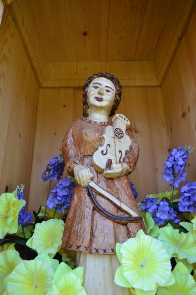 Drewniana rzeźba kobiety trzymającej skrzypce i smyczek, otoczona żółtymi i fioletowymi kwiatami, w drewnianej wnęce.