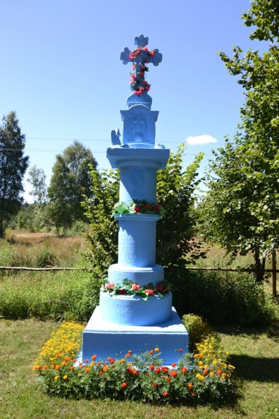 Niebieski kamienny krzyż na cokole, ozdobiony czerwonymi kwiatami, otoczony żółto-pomarańczowymi kwiatami, z drzewami i niebieskim niebem w tle.