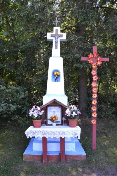 Biały kamienny krzyż z figurą Jezusa, otoczony kwiatami i mniejszym drewnianym krzyżem ozdobionym różowymi kwiatami, na tle zielonych drzew.