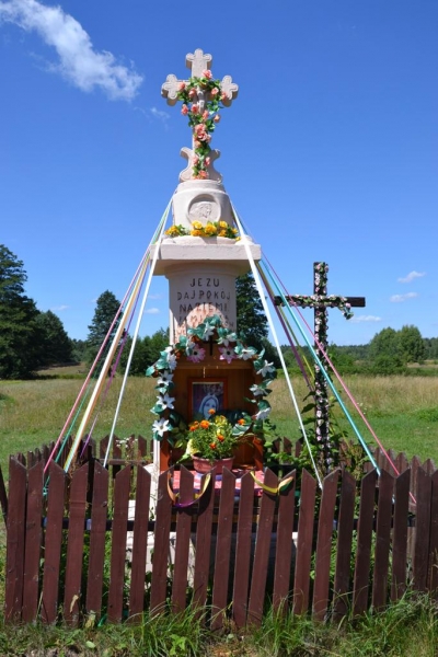 Kamienny biały krzyż z dekoracjami kwiatowymi i kolorowymi wstążkami, otoczony brązowym drewnianym płotkiem na tle zielonego pola i błękitnego nieba.