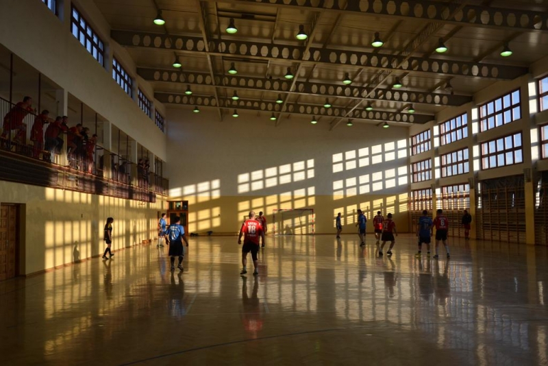 Hala sportowa z dużymi oknami, na parkiecie kilka osób w czerwonych i niebieskich strojach, widzowie na balkonie po lewej stronie, światło słoneczne tworzy wzory na ścianach i podłodze.