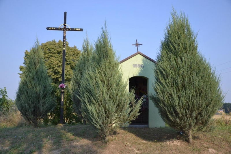 Mała zielona kapliczka z metalowym krzyżem na dachu i czarny metalowy krzyż z napisem, otoczona wysokimi zielonymi drzewami iglastymi na tle jasnoniebieskiego nieba.