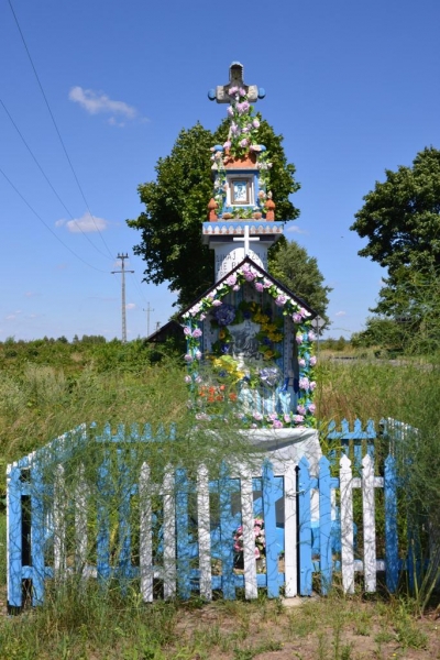 Kapliczka z krzyżem i kwiatami, otoczona niebiesko-białym drewnianym płotem, na tle zielonych drzew i niebieskiego nieba.