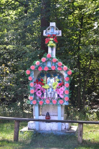 Biały drewniany krzyż z kolorowymi sztucznymi kwiatami w różowym, czerwonym i żółtym kolorze, ustawiony na betonowej podstawie na tle zielonego lasu.