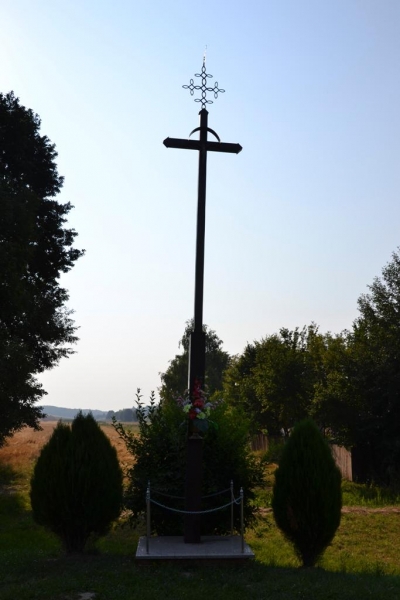 Wysoki, ciemny metalowy krzyż z ozdobnym zwieńczeniem, otoczony zielonymi krzewami i dwoma stożkowatymi drzewkami na tle jasnego nieba i drzew.