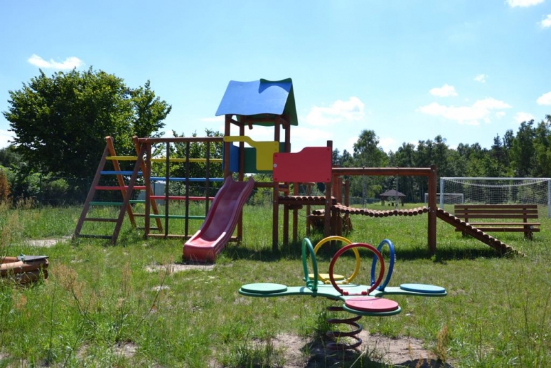 Plac zabaw na trawie z czerwonym zjeżdżalnią, drewnianą drabinką, mostkiem i huśtawką, zielone, niebieskie, czerwone i żółte elementy, w tle drzewa i niebieskie niebo.