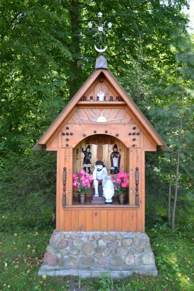 Kapliczka z drewna na kamiennej podstawie, z figurkami i różowymi kwiatami, otoczona zielonymi drzewami.