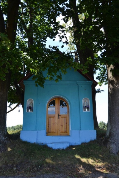 Niebieska kapliczka z drewnianymi drzwiami, osłonięta przez duże drzewa z zielonymi liśćmi, na tle trawy i nieba.