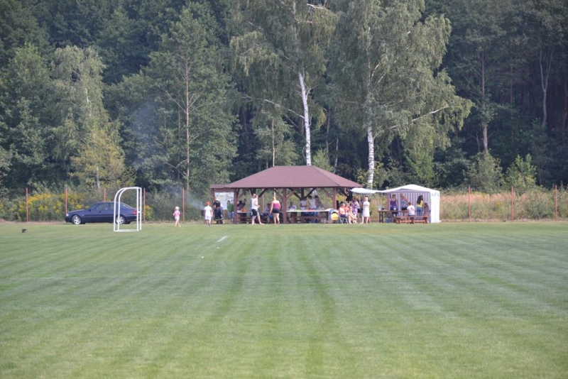 Boisko z zieloną trawą, na tle lasu, z altaną i grupą ludzi oraz stojącym samochodem po lewej stronie.