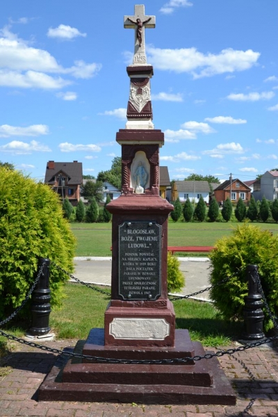 Kamienny krzyż z figurą Jezusa, cokół z napisem, mała figura Matki Boskiej, otoczenie zieleni i domów, niebieskie niebo z chmurami.