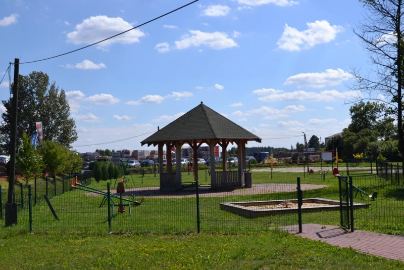 Plac zabaw z drewnianą altaną, piaskownicą i huśtawkami na tle zielonej trawy, drzew i niebieskiego nieba z chmurami.