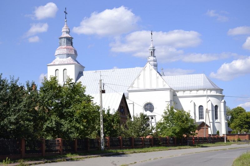 Biała świątynia z czerwonym dachem, wieża z krzyżem, otoczona zielonymi drzewami, pod niebieskim niebem z kilkoma białymi chmurami.