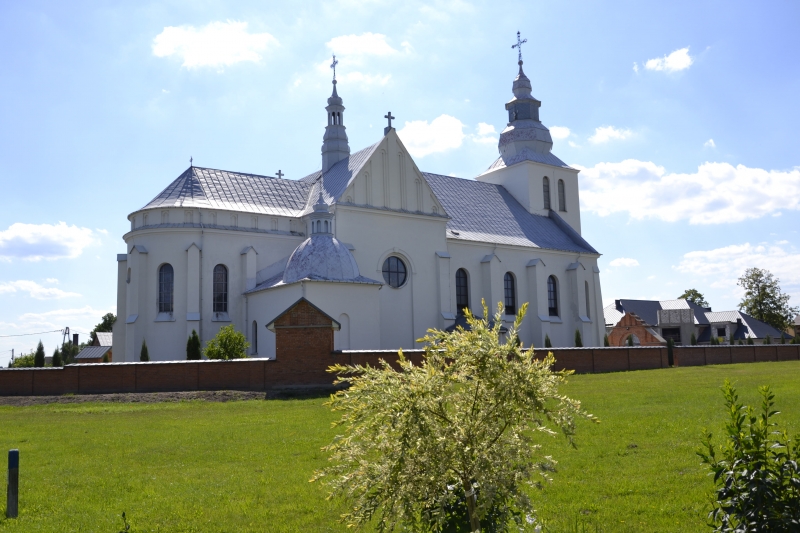 Biała barokowa świątynia z wieżami i krzyżami na dachu, otoczona ceglanym murem, na pierwszym planie zielona trawa i krzewy, niebieskie niebo z chmurami.