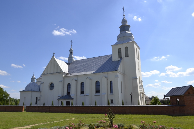 Biała neogotycka świątynia z metalowym dachem i wieżą z krzyżem, otoczona ceglanym murem, na zielonym trawniku pod błękitnym niebem z nielicznymi chmurami.