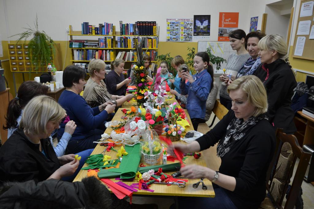 Grupa kobiet i dziewcząt siedzi oraz stoi przy długim stole z kolorowymi materiałami, kwiatami i narzędziami do rękodzieła w pomieszczeniu z regałami z książkami.