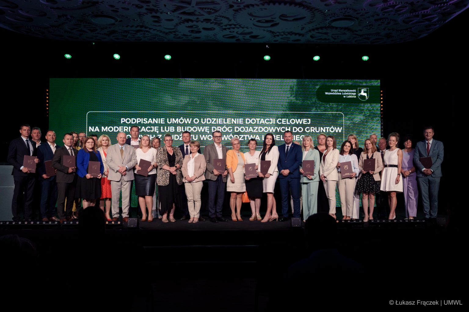 Grupa 27 osób w eleganckich strojach stoi na scenie przed zielonym ekranem z białym tekstem i logo Urzędu Marszałkowskiego Województwa Lubelskiego.