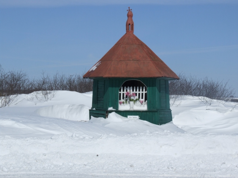 Niewielka zielona kapliczka z czerwoną, stożkową dachówką i oknem z białą kratą, z kwiatami w doniczce, otoczona śniegiem i krzewami pod jasnym, niebieskim niebem.