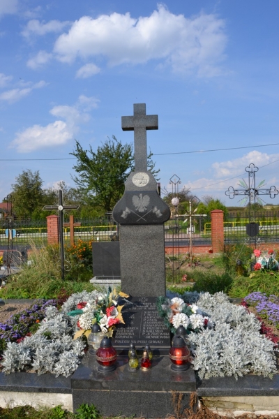 Szary kamienny nagrobek z krzyżem, ozdobiony białymi i fioletowymi kwiatami oraz kolorowymi zniczami, na tle niebieskiego nieba i zielonych drzew.