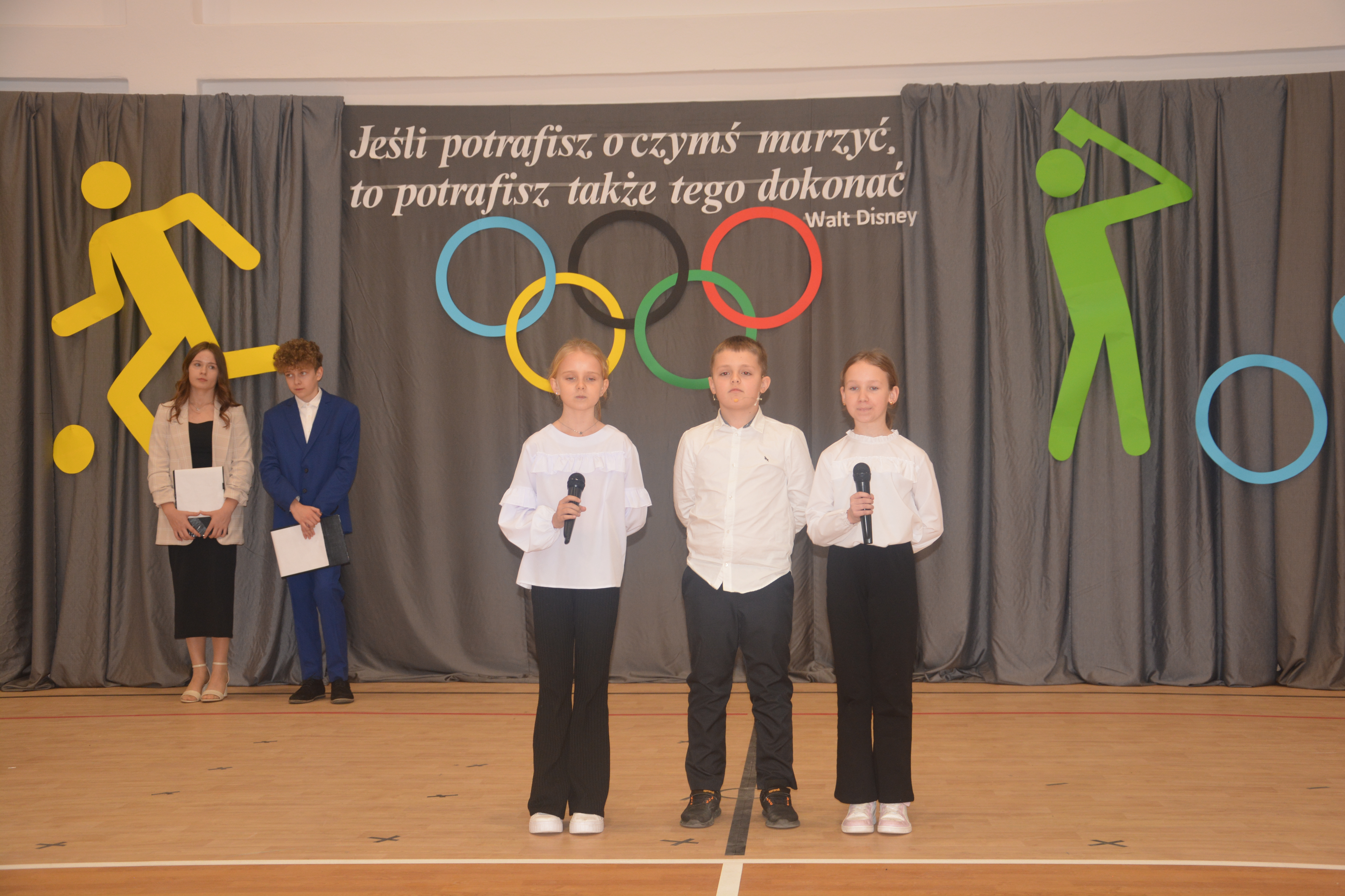 Trójka dzieci w białych bluzkach i ciemnych spodniach z mikrofonami na tle szarych zasłon z kolorowymi symbolami olimpijskimi i cytatem Walt Disney'a.