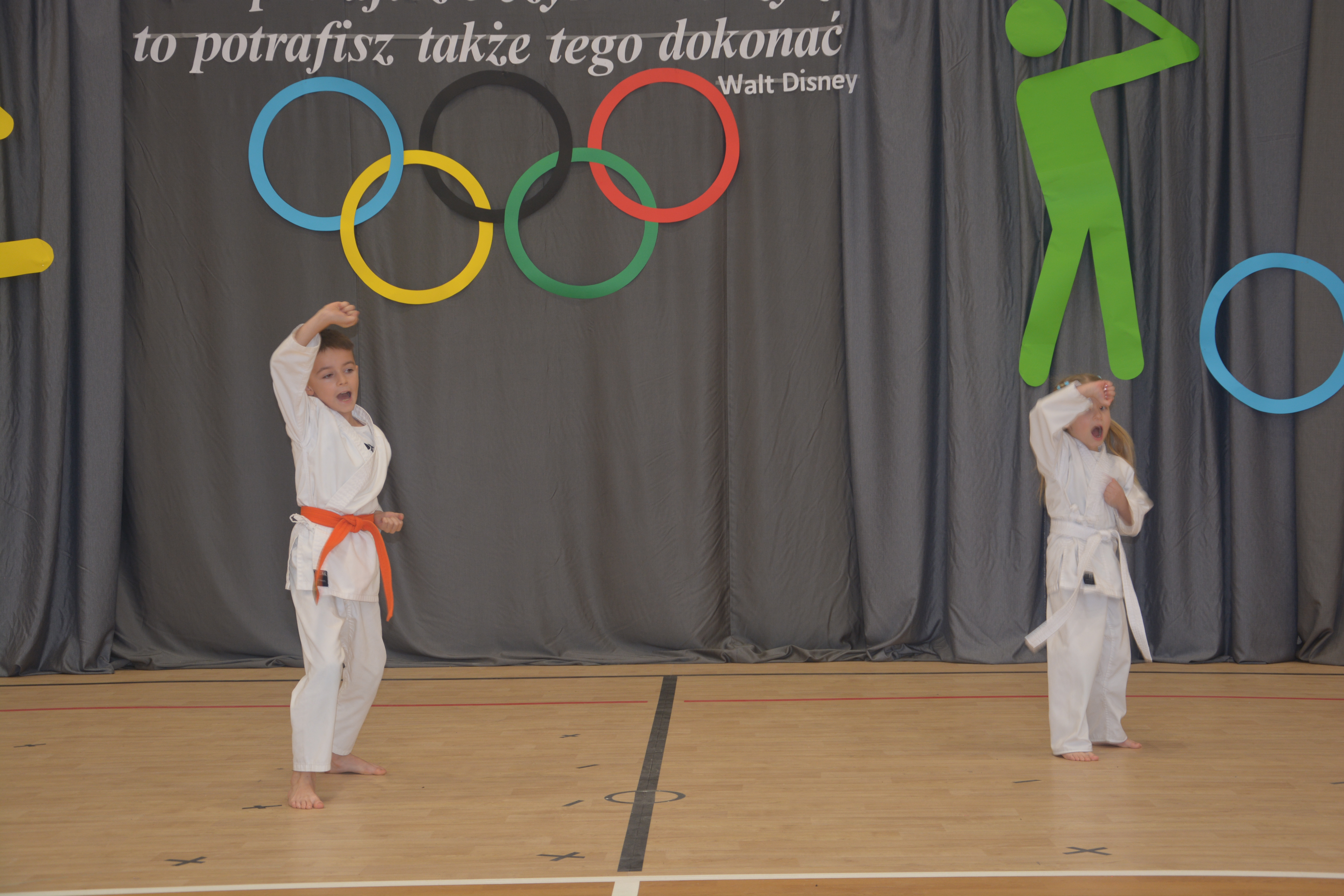 Dwójka dzieci w białych kimona, chłopiec z pomarańczowym pasem, dziewczynka z białym, ćwiczy karate na drewnianej podłodze, szare tło z kolorowymi olimpijskimi kołami i zielonym symbolem golfisty.