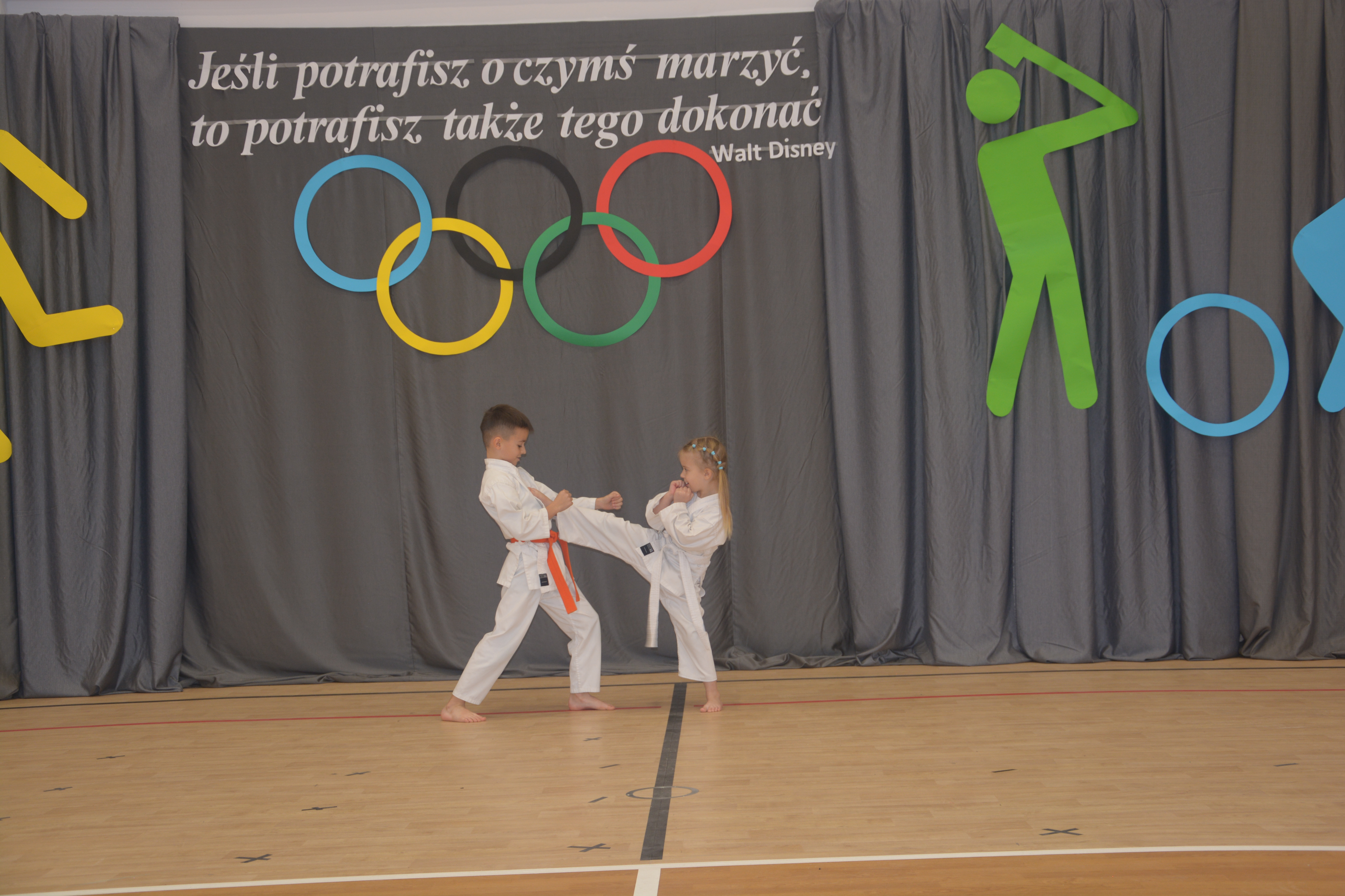 Dwoje dzieci w białych kimono ćwiczy karate na drewnianej sali gimnastycznej, w tle szare zasłony i kolorowe koła olimpijskie oraz sylwetki sportowców.