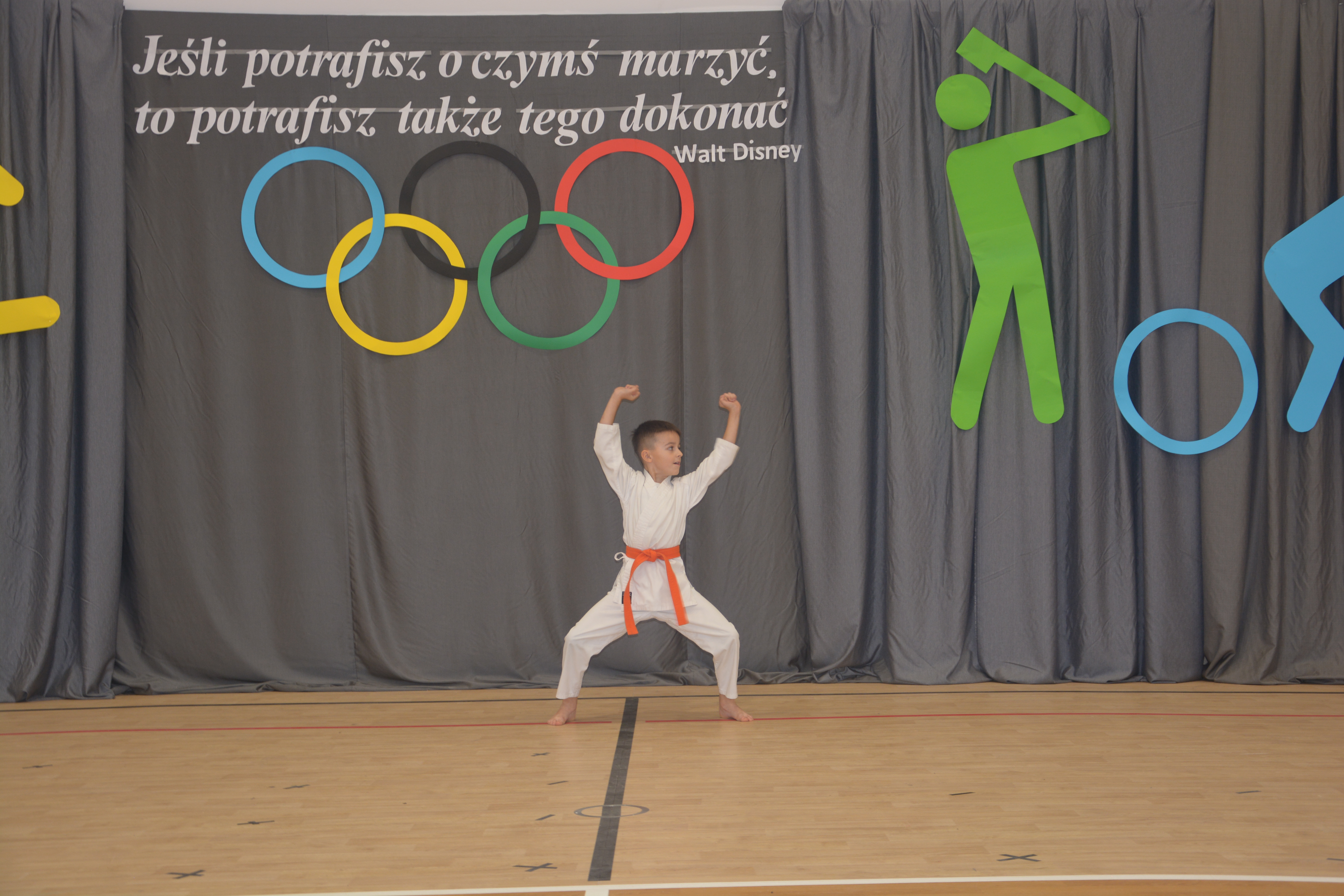 Chłopiec w białym kimonie z pomarańczowym pasem stoi w szerokim rozkroku na drewnianej podłodze, na szarym tle z kolorowymi symbolami olimpijskimi i cytatem w języku polskim.