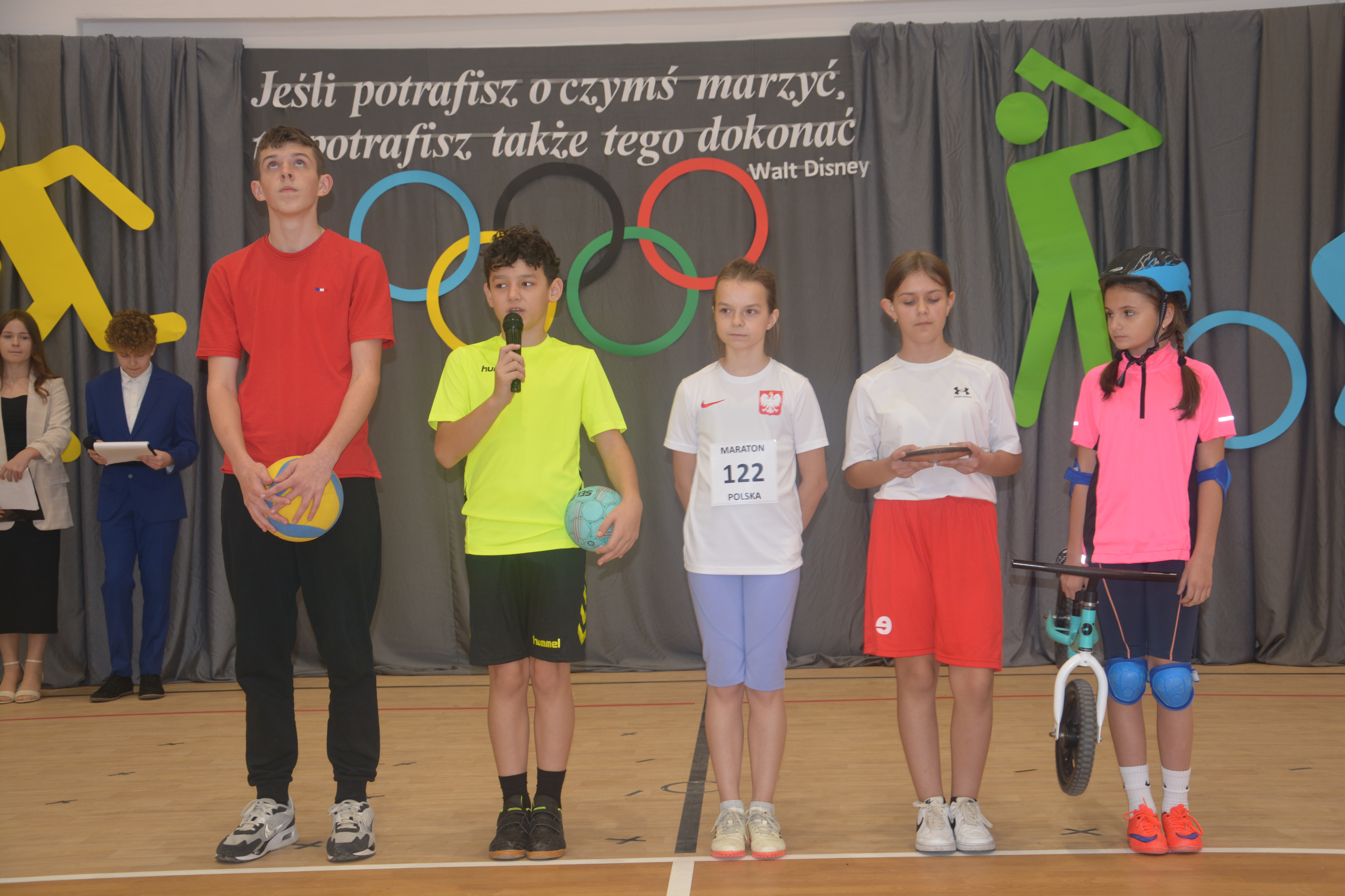 Pięcioro dzieci w sportowych strojach stoi na tle szarych zasłon z kolorowymi symbolami olimpijskimi i cytatem Walt Disneya. Jedno dziecko trzyma mikrofon, inne piłki, a jedno rowerek.