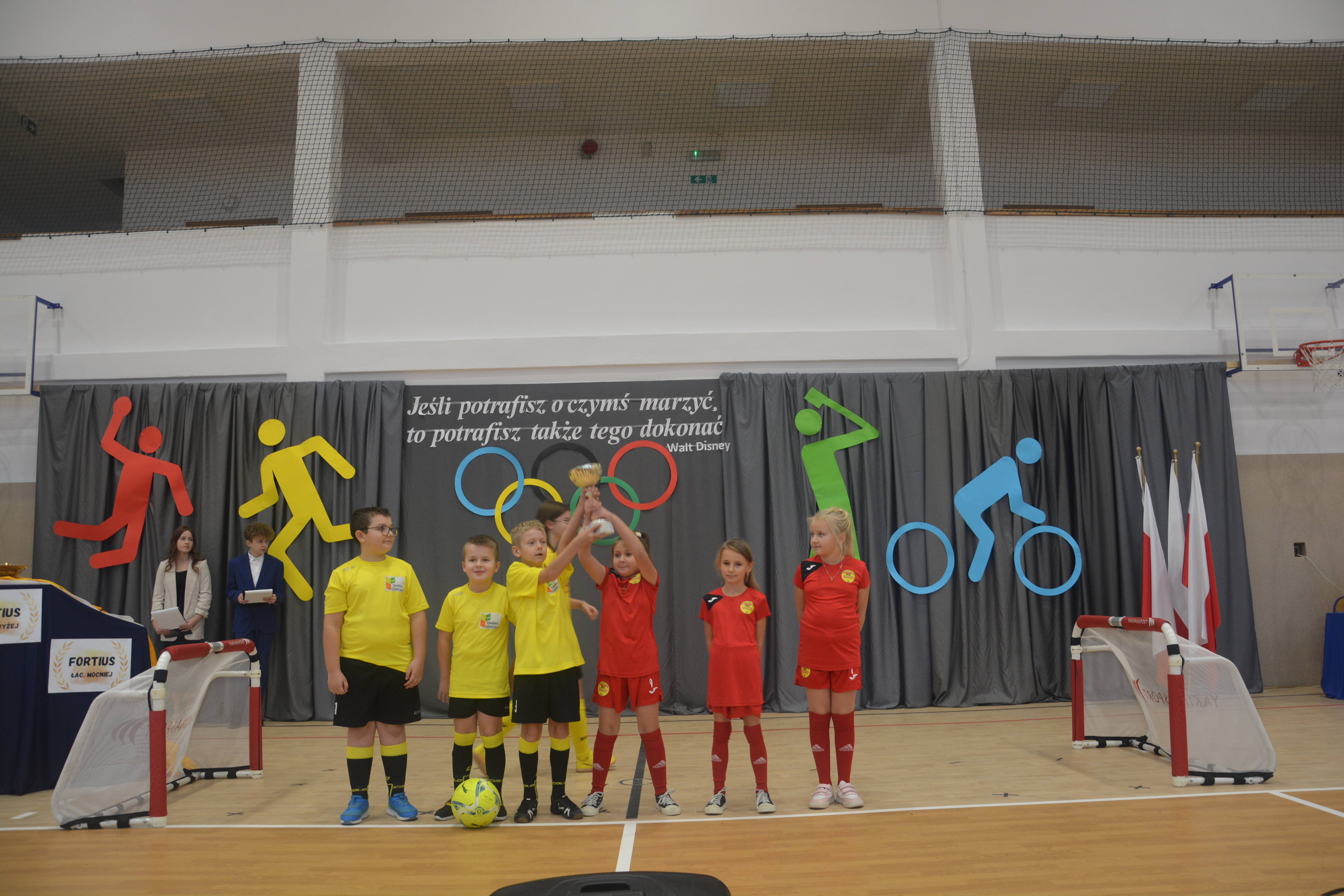 Grupa dzieci w żółtych i czerwonych strojach sportowych na hali z siatką i symbolami sportów na szarym tle, trzymająca puchar, obok dwie małe bramki do piłki nożnej.