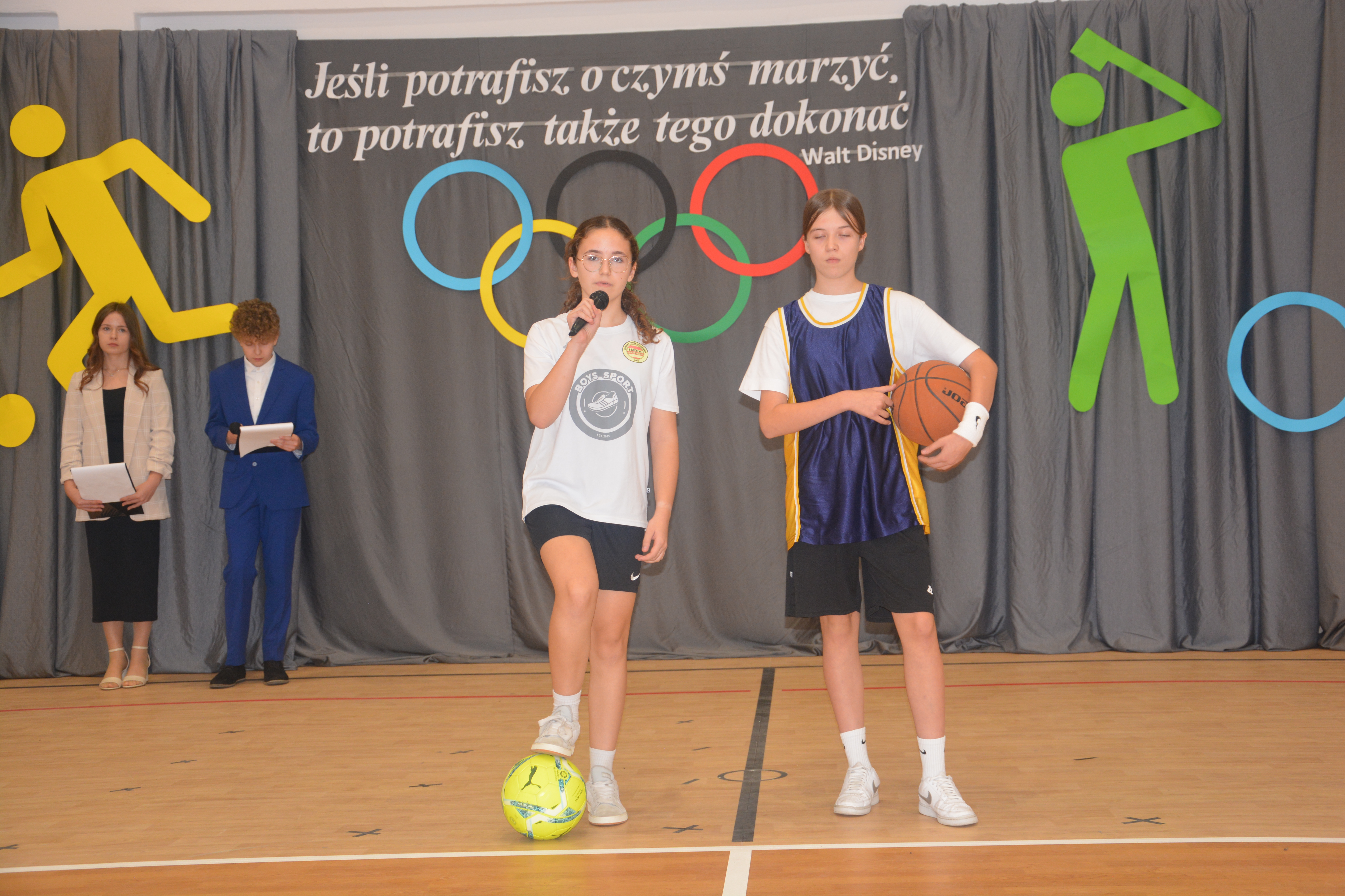 Cztery osoby na tle szarych zasłon z kolorowymi symbolami sportowymi i napisem, dwie dziewczyny na pierwszym planie w sportowych strojach, jedna trzyma mikrofon, druga piłkę do koszykówki.