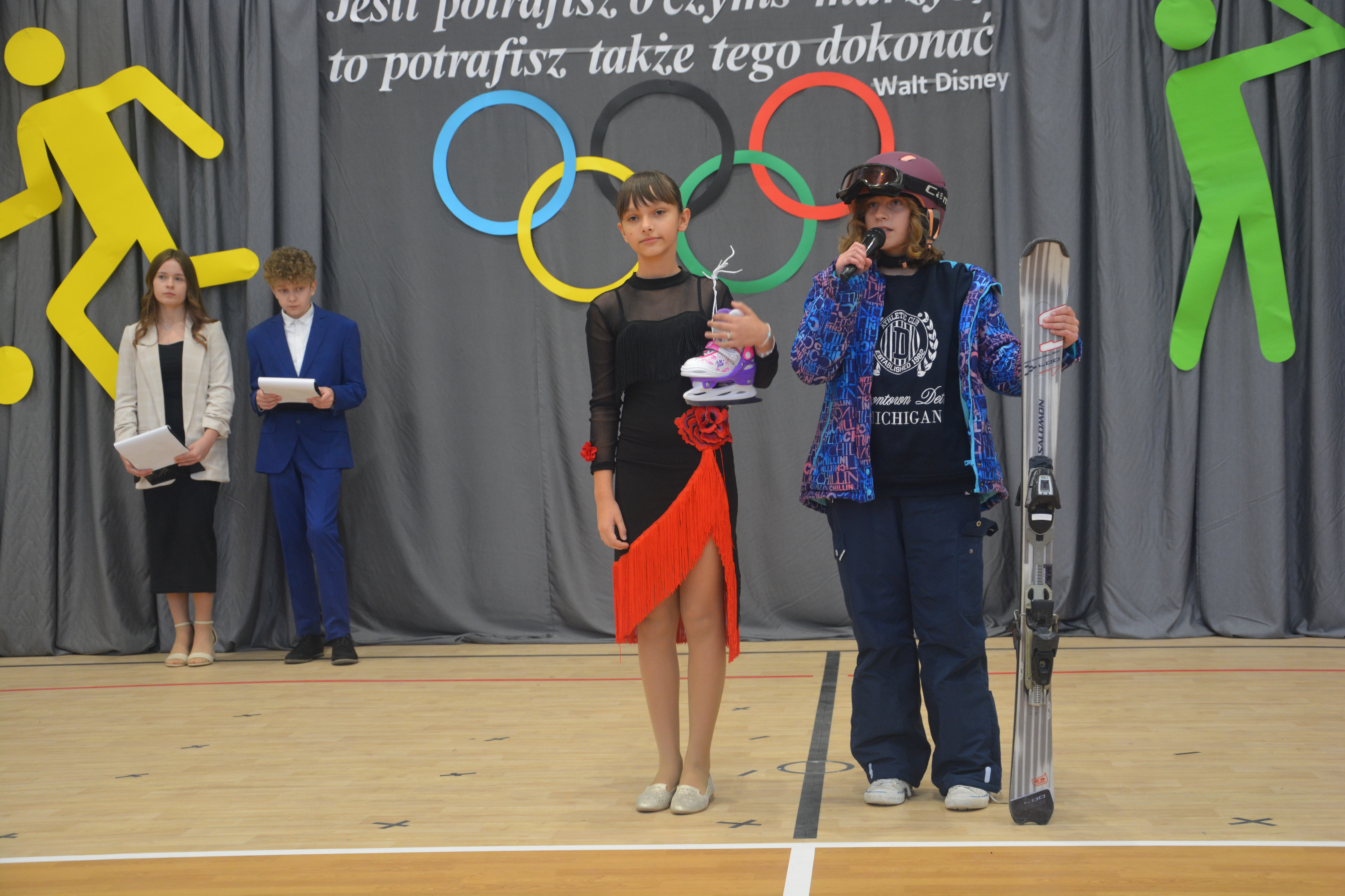 Cztery osoby na sali gimnastycznej, dwie z przodu trzymają łyżwy i nartę, za nimi dekoracje z kolorowymi olimpijskimi kołami i sylwetki sportowców.