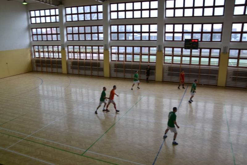 Sala gimnastyczna z drewnianą podłogą, dużymi oknami i pięcioma osobami w sportowych strojach, dwie osoby w zielonych koszulkach, jedna w pomarańczowej, jedna w czerwonej.