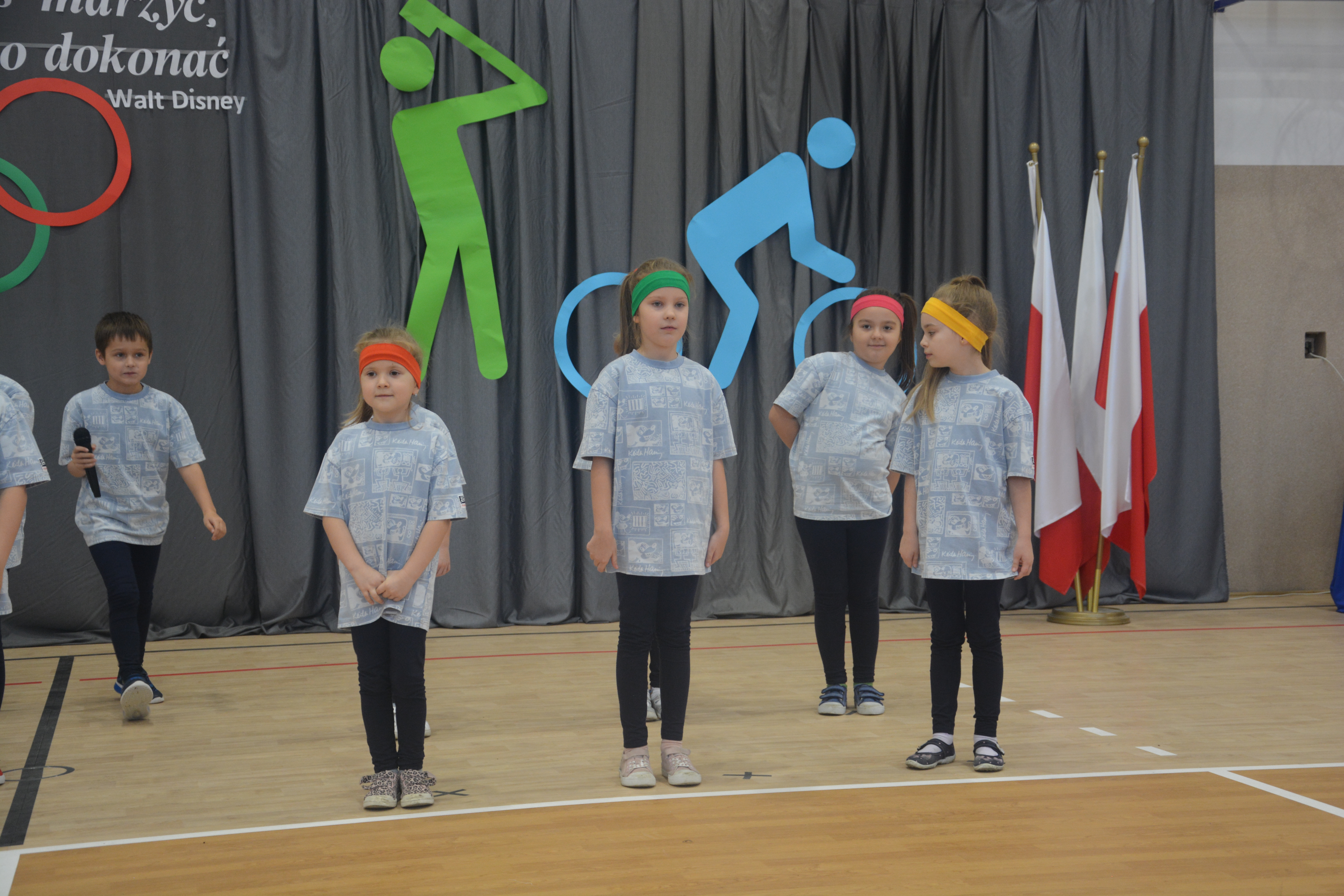 Grupa dzieci w szarych koszulkach i kolorowych opaskach na głowach stoi na drewnianej podłodze hali sportowej, w tle szare zasłony i polskie flagi.