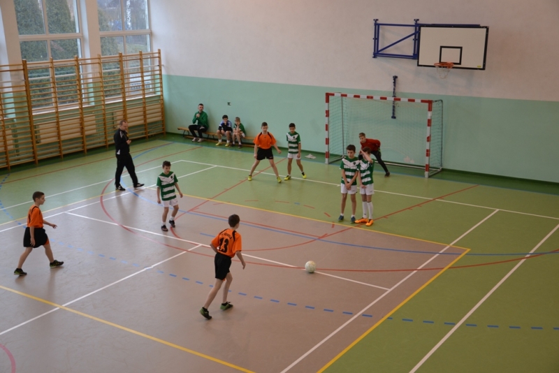 Dzieci w pomarańczowych i zielono-białych strojach na hali sportowej podczas gry w piłkę nożną, bramka i kosz na tle zielonych ścian i dużych okien.