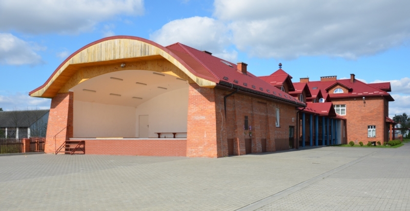 Ceglana budowla z dużą, półokrągłą, drewnianą sceną i czerwonym dachem, otoczona szarą kostką brukową pod niebieskim niebem z chmurami.