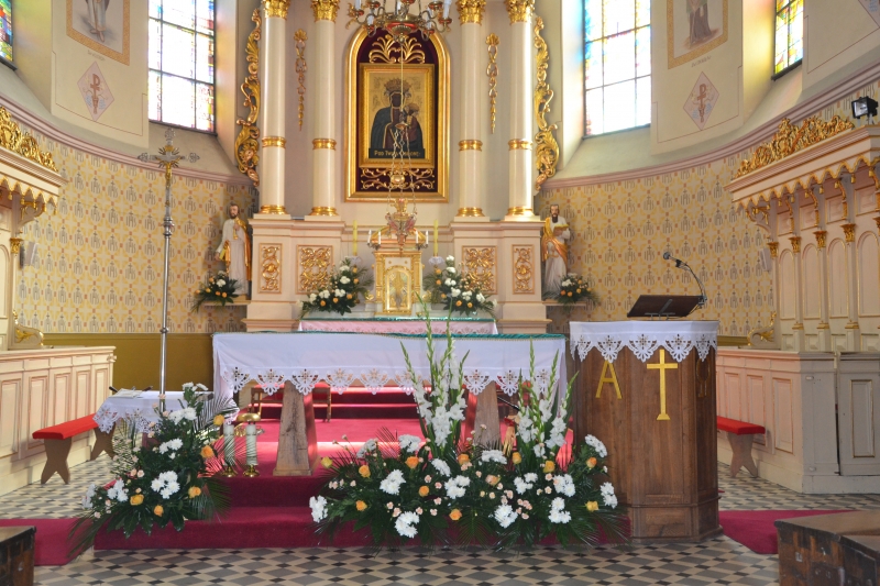 Wnętrze kościoła z ołtarzem, białym obrusem, bukietami białych i pomarańczowych kwiatów, drewnianą amboną z białą koronką i złotym krzyżem, ornamentami w kolorze złotym i czerwonym dywanem.