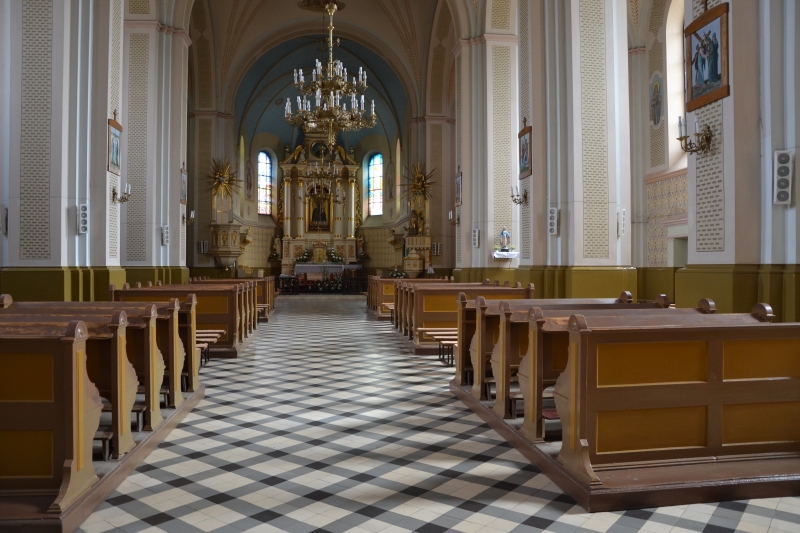 Wnętrze kościoła z drewnianymi ławkami, jasnobeżowymi ścianami, dużym żyrandolem i ołtarzem z ozdobami oraz dwoma witrażowymi oknami.