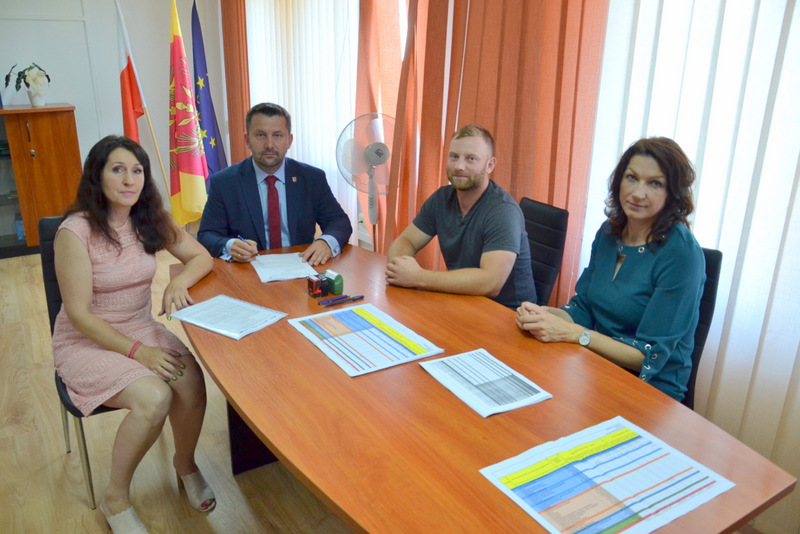 Cztery osoby siedzące przy drewnianym stole, na stole dokumenty z kolorowymi wykresami, w tle pomarańczowe i białe zasłony oraz trzy flagi.