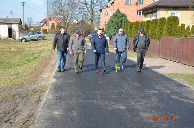 Pięciu mężczyzn w kurtkach idzie po świeżo położonej asfaltowej drodze wśród domów jednorodzinnych, zieleń i samochód po lewej stronie.