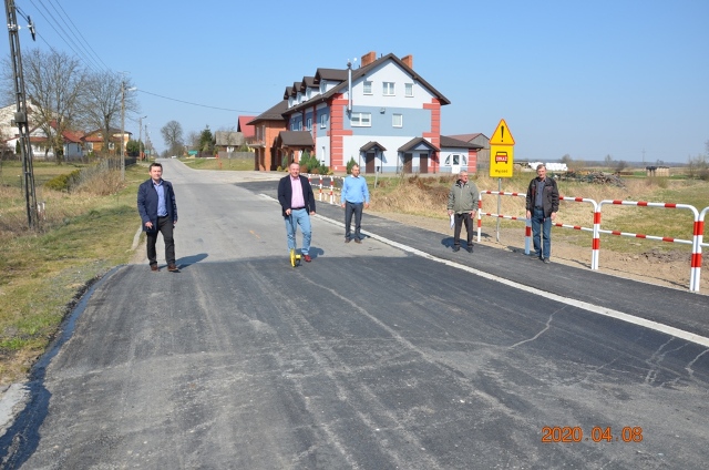 Pięć osób stoi na nowo wyasfaltowanej szarej drodze przy jasnym, bezchmurnym niebie, w tle jasnoniebieski dom z czerwonymi elementami. Po prawej stronie biało-czerwone barierki i znaki drogowe.