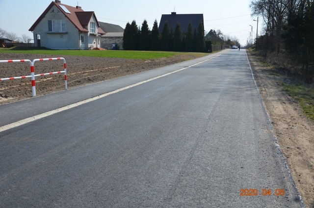 Asfaltowa droga z białą linią krawędziową, po lewej stronie dom z czerwonym dachem, pole uprawne i zielony trawnik, po prawej stronie drzewa i pobocze z piasku.