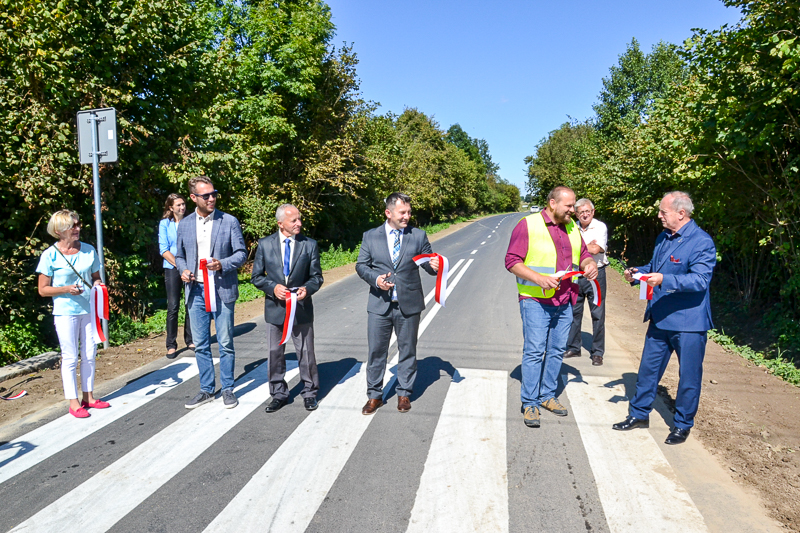 Siedem osób stoi na przejściu dla pieszych na nowej drodze, trzymają białoczerwone wstążki i nożyczki, w tle zielone drzewa i jasne niebo.